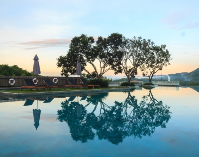 Tranquil swimming pool at sunset, reflecting trees and umbrellas, surrounded by lush greenery and mountains in the background.