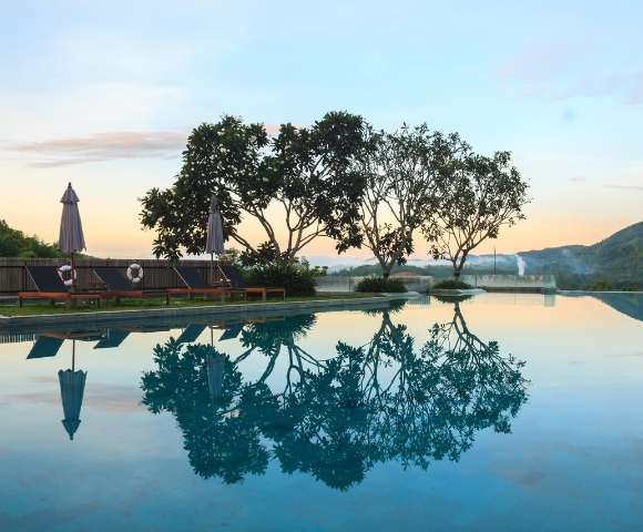 Tranquil swimming pool at sunset, reflecting trees and umbrellas, surrounded by lush greenery and mountains in the background.