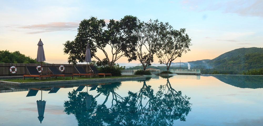 Tranquil swimming pool at sunset, reflecting trees and umbrellas, surrounded by lush greenery and mountains in the background.