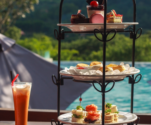 A tiered stand displays an assortment of pastries and sandwiches, with a refreshing drink beside it, set against a scenic background.
