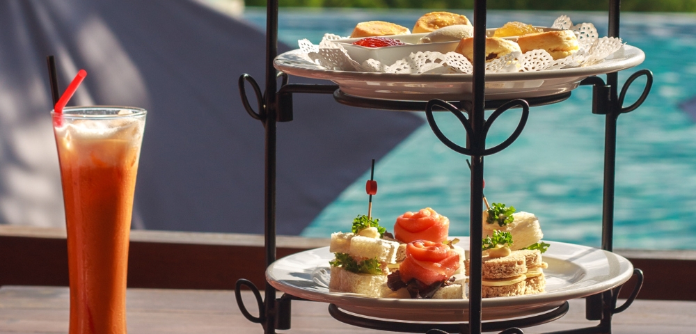 A tiered stand displays an assortment of pastries and sandwiches, with a refreshing drink beside it, set against a scenic background.