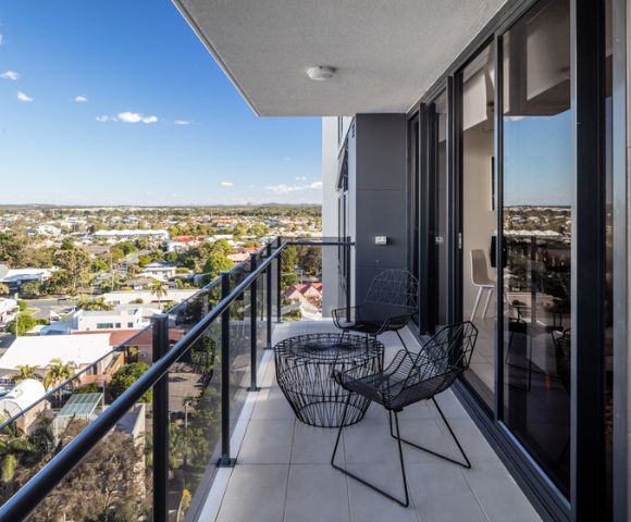A modern balcony with two black wire chairs and a round table, overlooking a sunny suburban landscape under a clear blue sky.