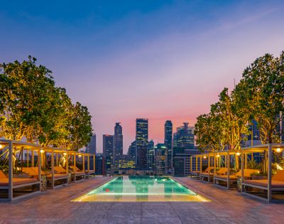 Rooftop poolside view with illuminated trees and loungers against a stunning city skyline at sunset.