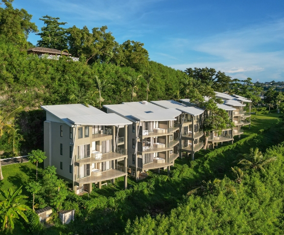 Modern multi-story buildings on a lush hillside, surrounded by greenery and palm trees, under a clear blue sky.