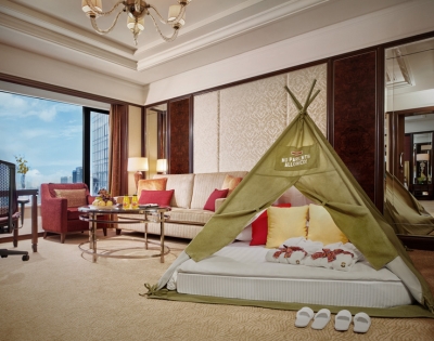 A luxury hotel room featuring a children's tent on a plush bed, colorful cushions, and a cozy seating area with city views.