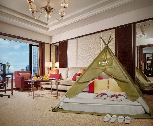 A luxury hotel room featuring a children's tent on a plush bed, colorful cushions, and a cozy seating area with city views.