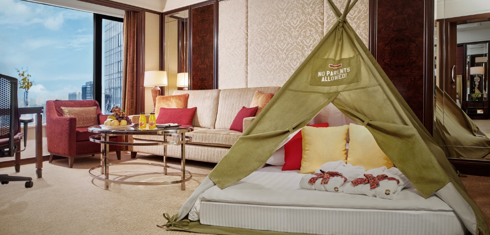 A luxury hotel room featuring a children's tent on a plush bed, colorful cushions, and a cozy seating area with city views.