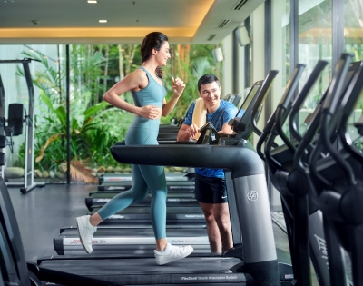A woman jogs on a treadmill in a bright gym, while a man beside her offers guidance. Lush greenery is visible through large windows.