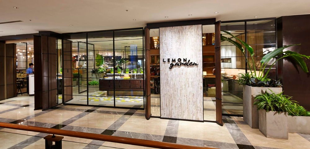 Entrance of Lemon Garden restaurant, featuring a stylish sign and glass walls, with a view of lush greenery inside.
