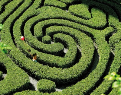 Aerial view of a child navigating through a green hedged maze, holding a red balloon, surrounded by lush greenery.