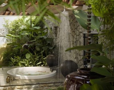 A serene outdoor bathing area with a circular tub, surrounded by lush greenery and a shower cascading from above.