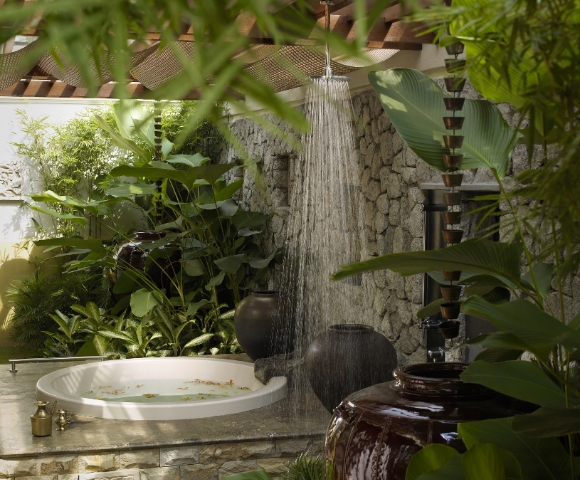 A serene outdoor bathing area with a circular tub, surrounded by lush greenery and a shower cascading from above.