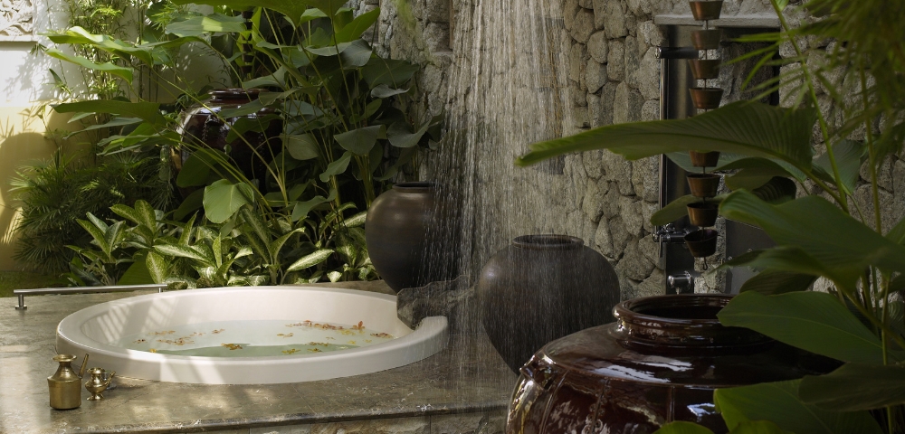 A serene outdoor bathing area with a circular tub, surrounded by lush greenery and a shower cascading from above.