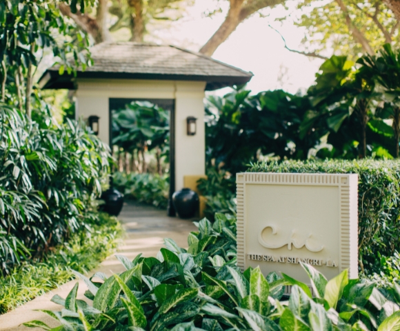 A serene entrance to a wellness retreat, framed by lush greenery, with a sign displaying 