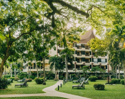 A lush green garden with lounge chairs and a path leading to a modern building surrounded by trees and tropical plants.