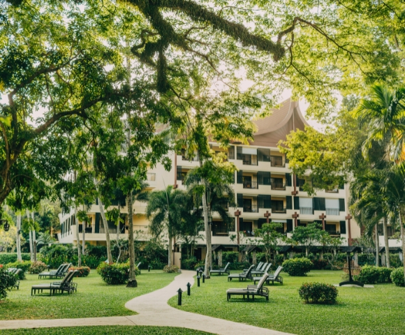 A lush green garden with lounge chairs and a path leading to a modern building surrounded by trees and tropical plants.
