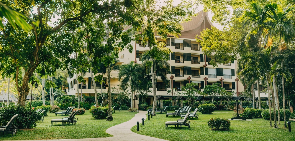A lush green garden with lounge chairs and a path leading to a modern building surrounded by trees and tropical plants.