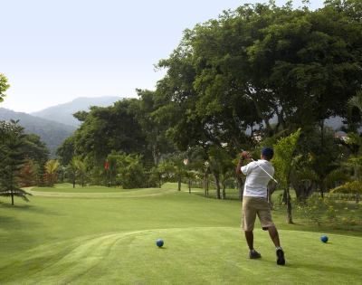 A golfer swings on a lush green course, surrounded by trees and mountains under a clear sky. Two blue tees mark the starting point.