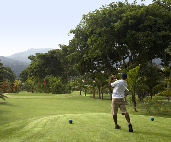 A golfer swings on a lush green course, surrounded by trees and mountains under a clear sky. Two blue tees mark the starting point.