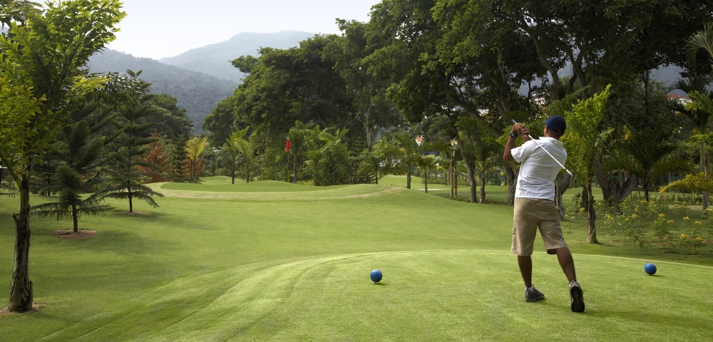 A golfer swings on a lush green course, surrounded by trees and mountains under a clear sky. Two blue tees mark the starting point.