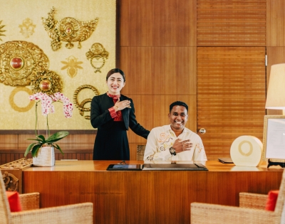 A receptionist and staff member stand behind a wooden desk adorned with orchids and decorative art in a luxurious lobby setting.