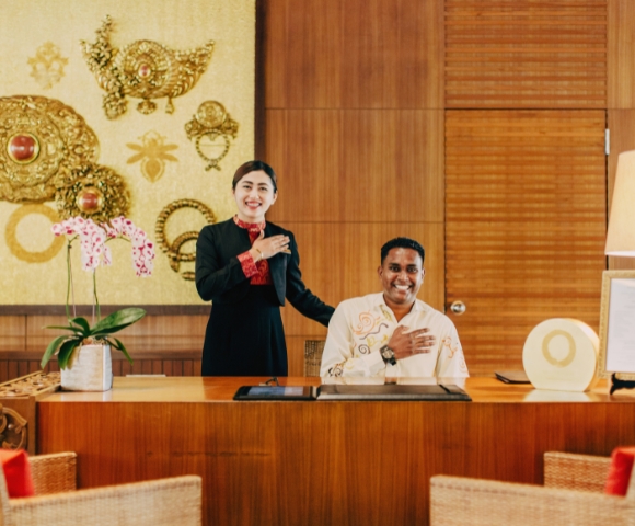 A receptionist and staff member stand behind a wooden desk adorned with orchids and decorative art in a luxurious lobby setting.