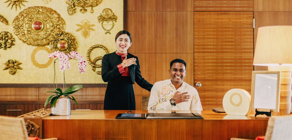 A receptionist and staff member stand behind a wooden desk adorned with orchids and decorative art in a luxurious lobby setting.