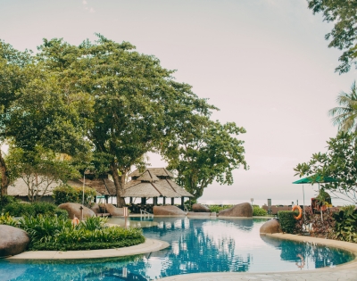 A serene resort pool surrounded by lush greenery and tropical trees, featuring a thatched roof pavilion and gentle curves along the water.