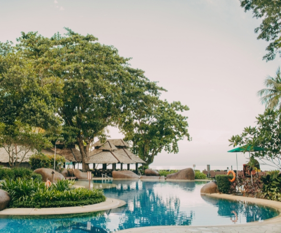 A serene resort pool surrounded by lush greenery and tropical trees, featuring a thatched roof pavilion and gentle curves along the water.