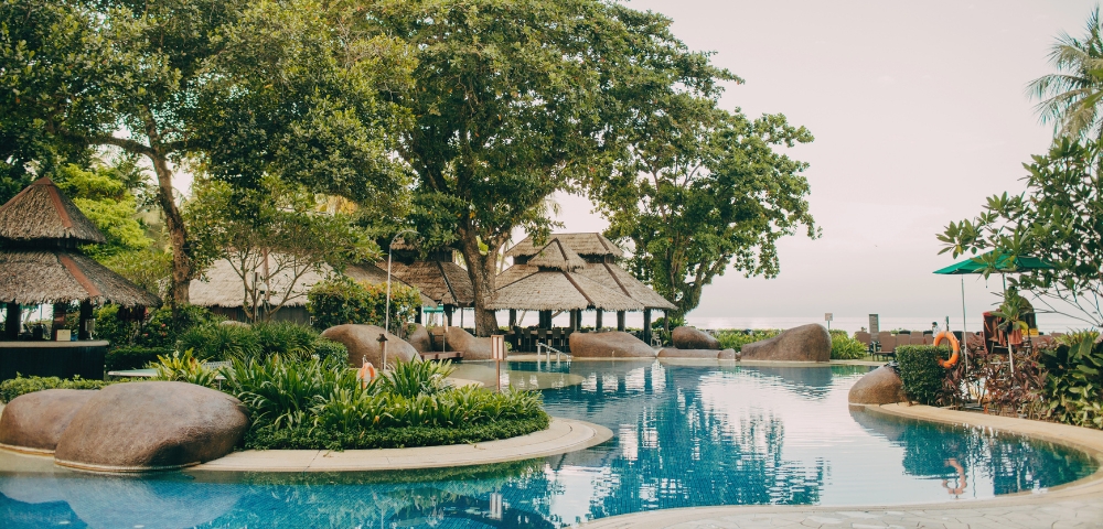 A serene resort pool surrounded by lush greenery and tropical trees, featuring a thatched roof pavilion and gentle curves along the water.
