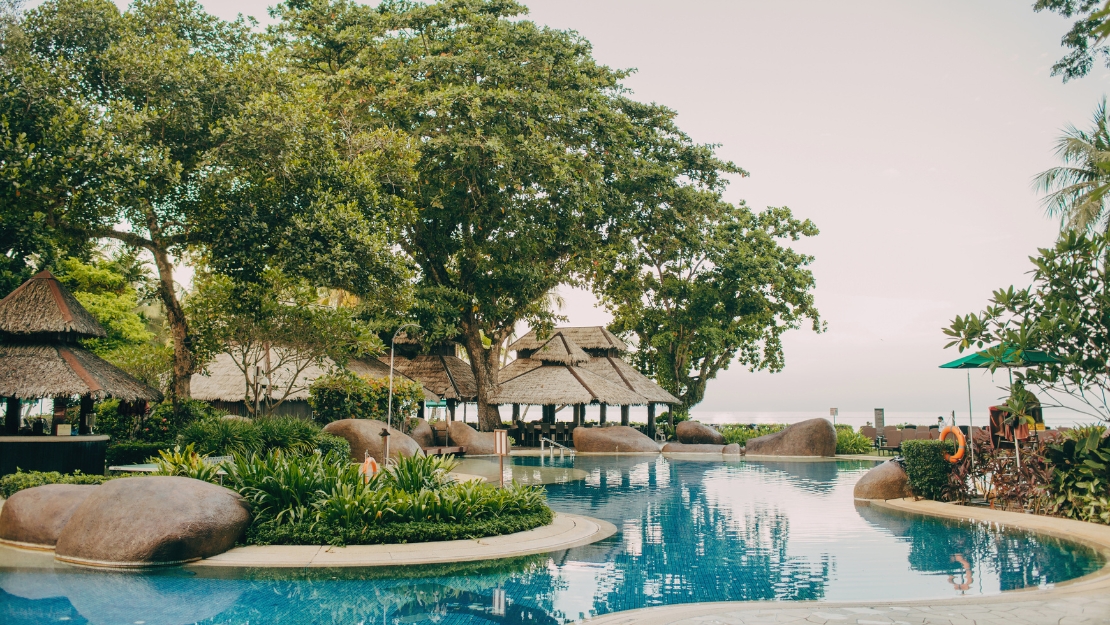 A serene resort pool surrounded by lush greenery and tropical trees, featuring a thatched roof pavilion and gentle curves along the water.