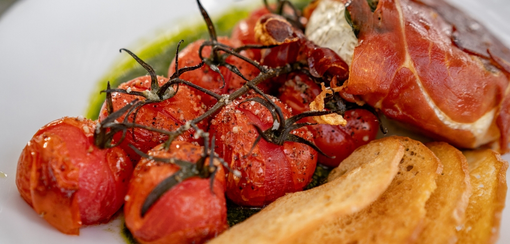 A plate featuring roasted cherry tomatoes on the vine, wrapped in prosciutto, accompanied by crispy bread and drizzled with green sauce.