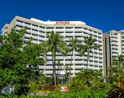 Rydges hotel building surrounded by palm trees and lush greenery under a clear blue sky.