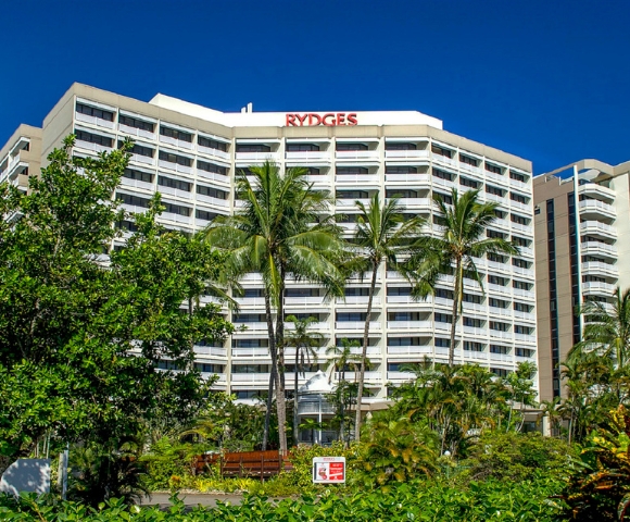 Rydges hotel building surrounded by palm trees and lush greenery under a clear blue sky.