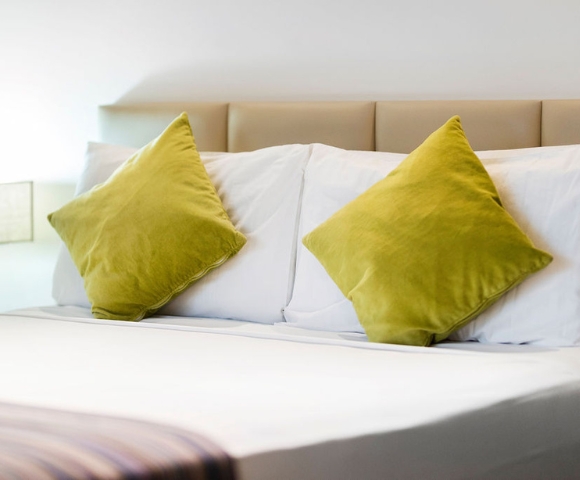 A neatly made bed with two vibrant green pillows against a light beige headboard and a soft striped blanket at the foot.
