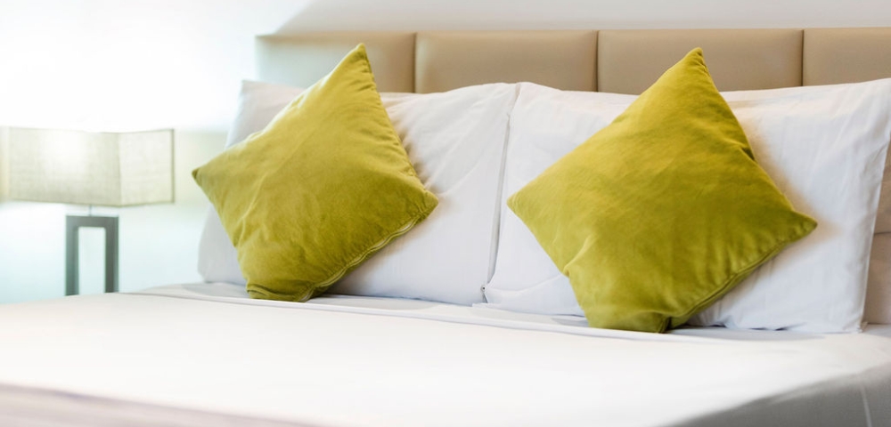 A neatly made bed with two vibrant green pillows against a light beige headboard and a soft striped blanket at the foot.