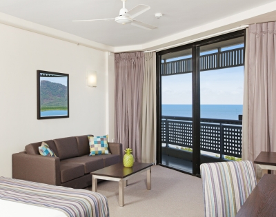 Cozy hotel room featuring a sofa, table, and large windows with ocean views, complemented by light curtains and a tropical decor.