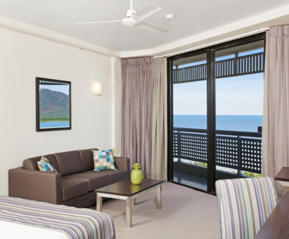 Cozy hotel room featuring a sofa, table, and large windows with ocean views, complemented by light curtains and a tropical decor.