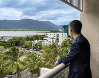 A man in a suit stands on a balcony, gazing out at a scenic view of mountains, trees, and a calm bay.