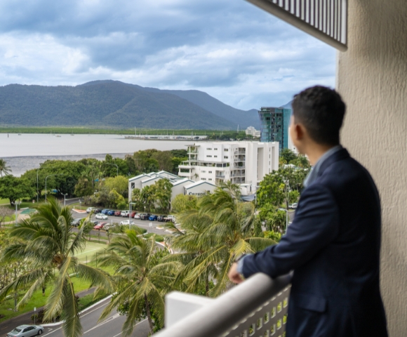 A man in a suit stands on a balcony, gazing out at a scenic view of mountains, trees, and a calm bay.