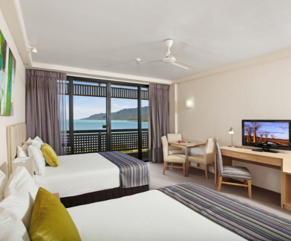 A bright hotel room featuring two beds, a desk, and a TV, with large windows showcasing a picturesque sea view.