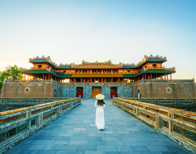 A woman in traditional attire walks towards a grand, historic temple, under a clear blue sky, surrounded by lush greenery.