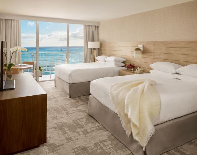 Modern hotel room with two beds, ocean view through large windows, wooden accents, and stylish decor.