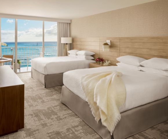 Modern hotel room with two beds, ocean view through large windows, wooden accents, and stylish decor.