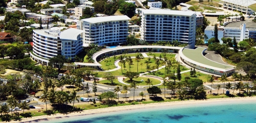 Aerial view of a modern hotel complex with palm trees and landscaped gardens by the beach, surrounded by urban buildings.