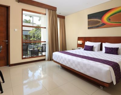 Modern hotel room featuring a large double bed, wooden accents, and a window view of a balcony with greenery. Colorful abstract art decorates the wall.