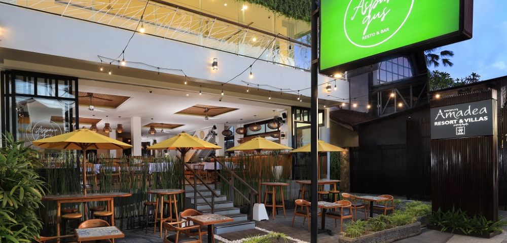 Outdoor seating of Alspana Resto & Bar featuring wooden tables, yellow umbrellas, and lush greenery, set in a modern building.