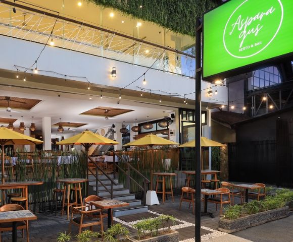 Outdoor seating of Alspana Resto & Bar featuring wooden tables, yellow umbrellas, and lush greenery, set in a modern building.