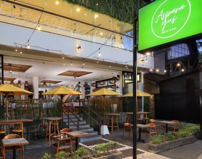 Outdoor seating of Alspana Resto & Bar featuring wooden tables, yellow umbrellas, and lush greenery, set in a modern building.