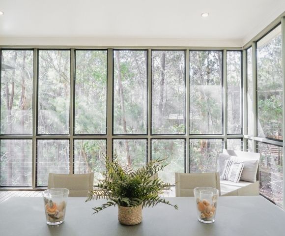 A bright sunroom with large windows overlooking greenery, featuring a table with a potted plant and glasses on it.